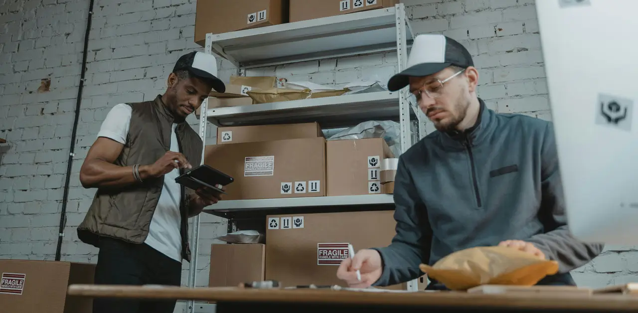 In A Warehouse, Two Men Are Busy Working Among Boxes And Computers, Demonstrating Teamwork And Productivity