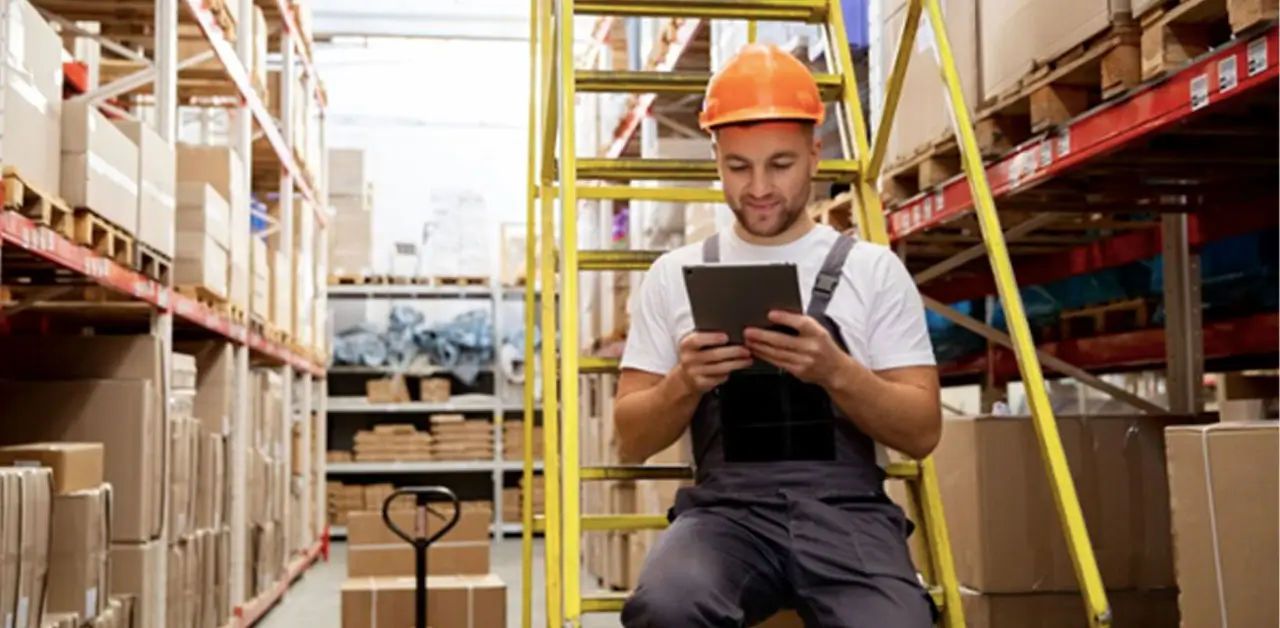 A Man In Overalls And A Hard Hat Sits On A Ladder In A Warehouse, With Various Tools And Shelves Around Him