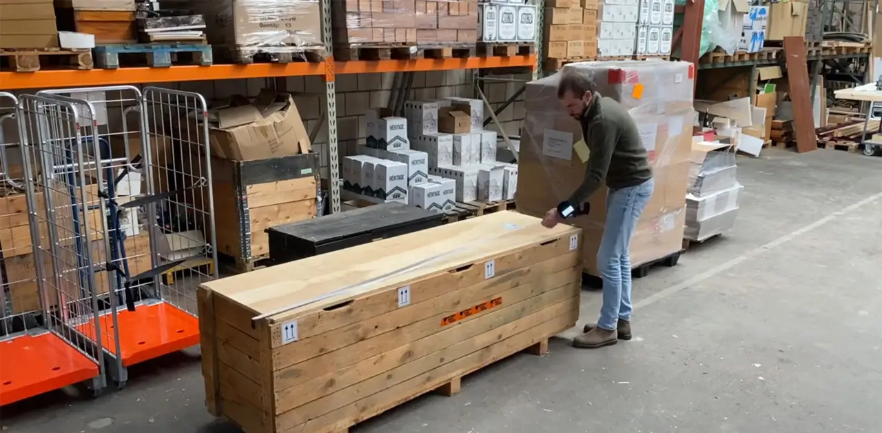 A Man Is Pushing A Large Wooden Crate Across The Floor Of A Warehouse, Surrounded By Shelves And Other Storage Items