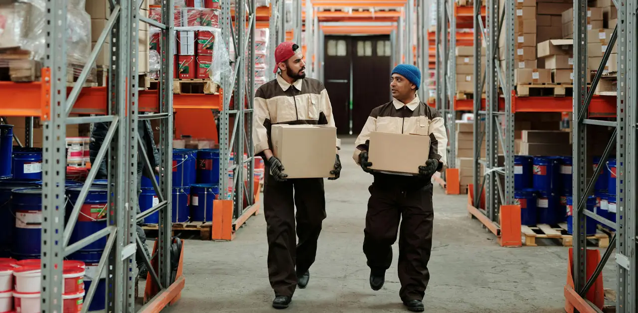 Two Warehouse Workers Moving Boxes Together, Highlighting Their Cooperation And The Bustling Atmosphere Of The Facility