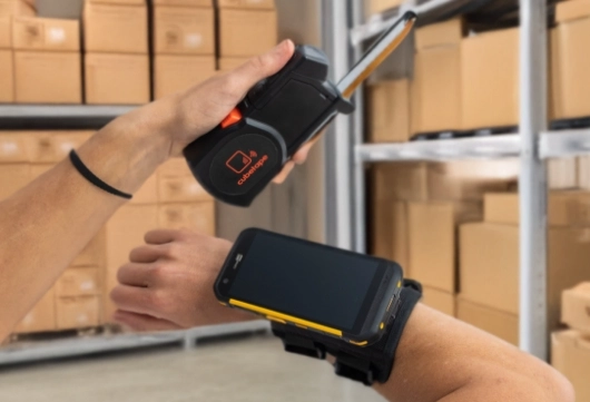 In A Warehouse, An Individual Holds A Cell Phone, Focused On The Device Amidst Rows Of Storage Shelves